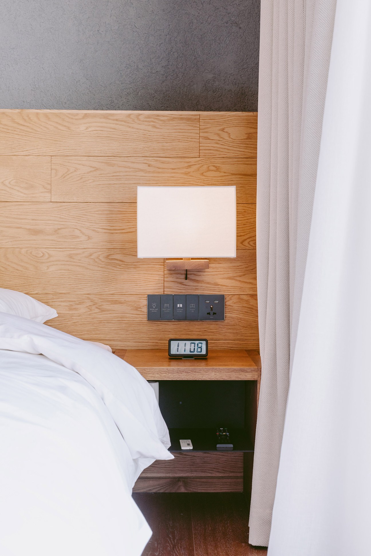 Muji Hotel bedside with light wood headboard, wall-mounted lamp, control switches and digital clock.
