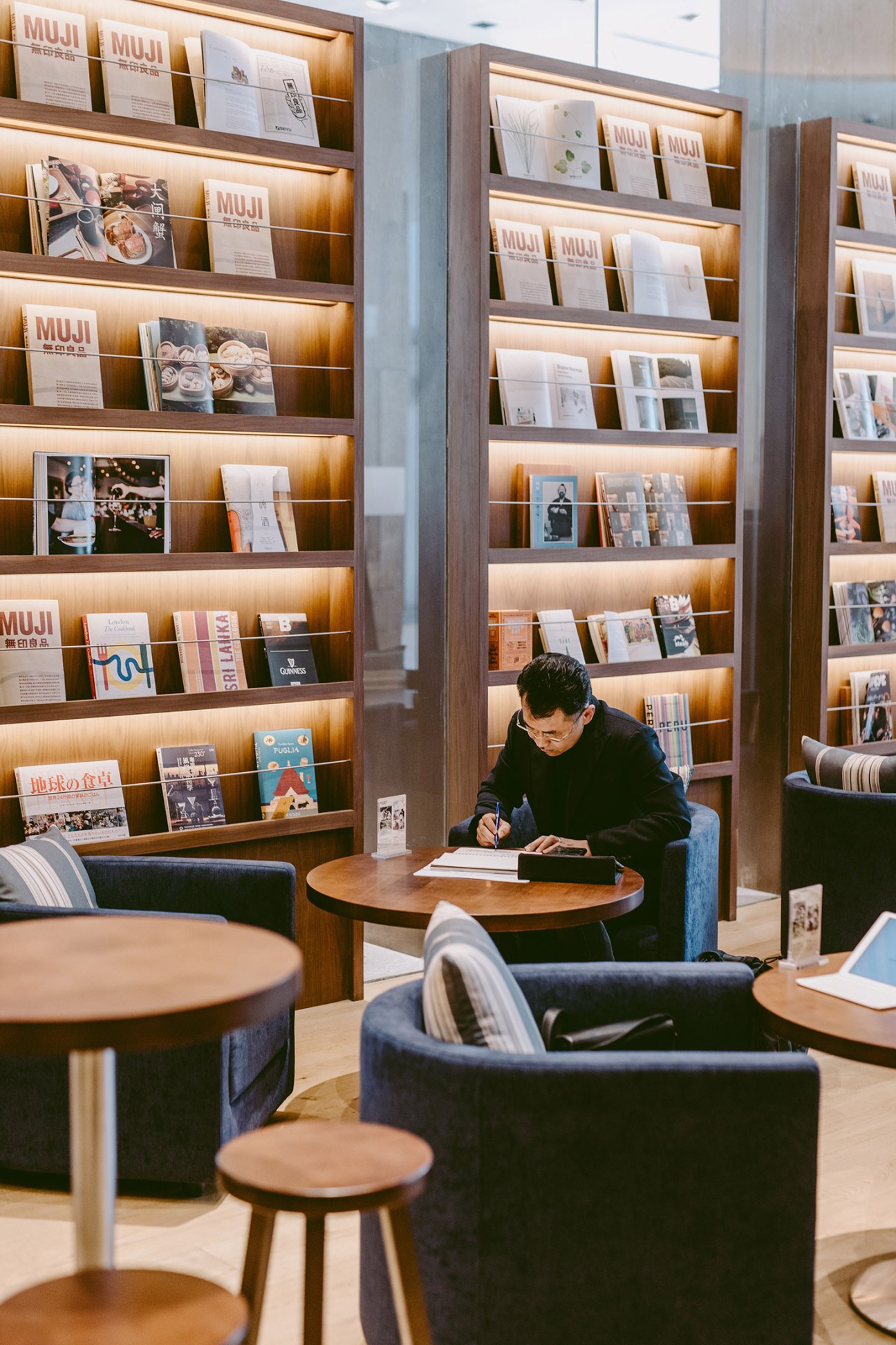 Person reading at wooden table in Muji Hotel lobby with illuminated bookshelves behind.