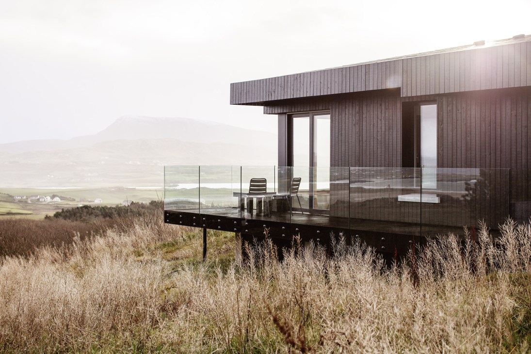Dark larch-clad Breac House with floor-to-ceiling windows overlooking heather landscape and Sheephaven Bay in Donegal, Ireland.