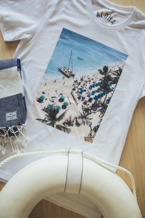 White t-shirt with beach scene print featuring sailboat, palm trees and blue umbrellas displayed with a striped scarf and white swim ring.