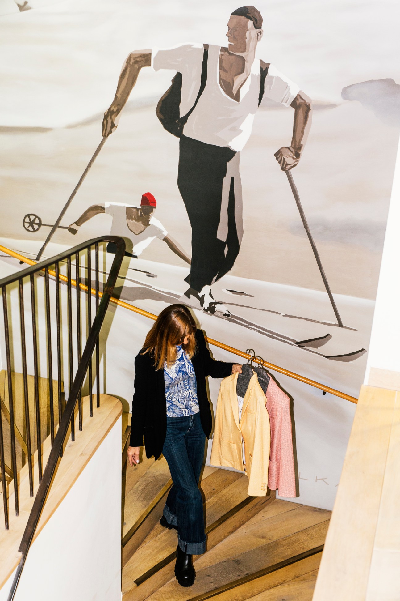 Woman on wooden stairs holding clothing items with large mural of skiers on wall behind