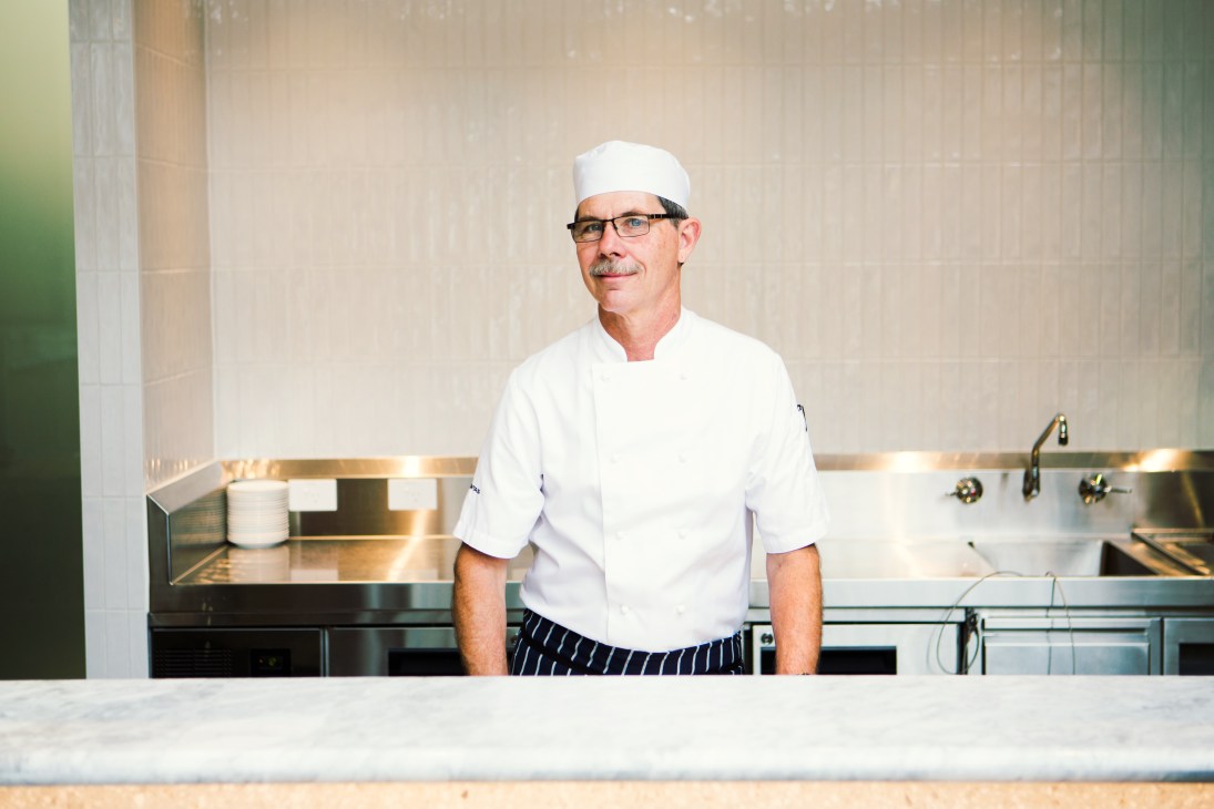 Chef in white uniform and hat standing behind a kitchen counter in a modern kitchen.
