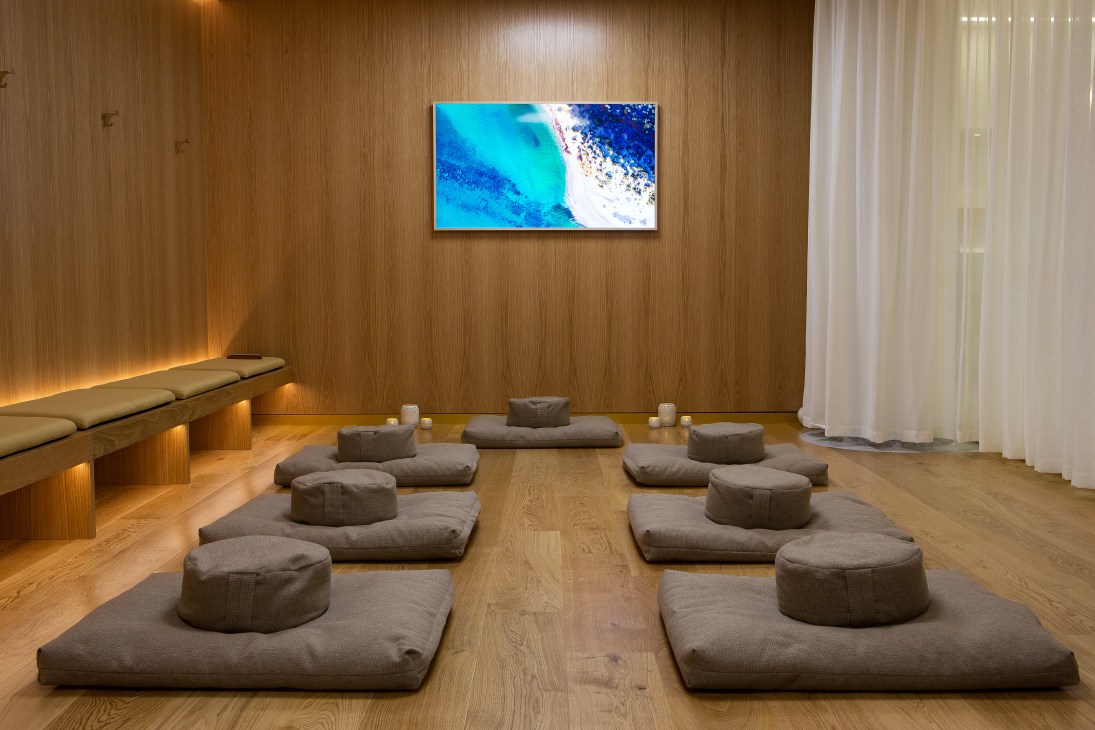 A minimalist wellness studio with wooden walls, gray cushioned floor mats, ambient lighting, and an aerial coastal landscape displayed on a wall-mounted screen.