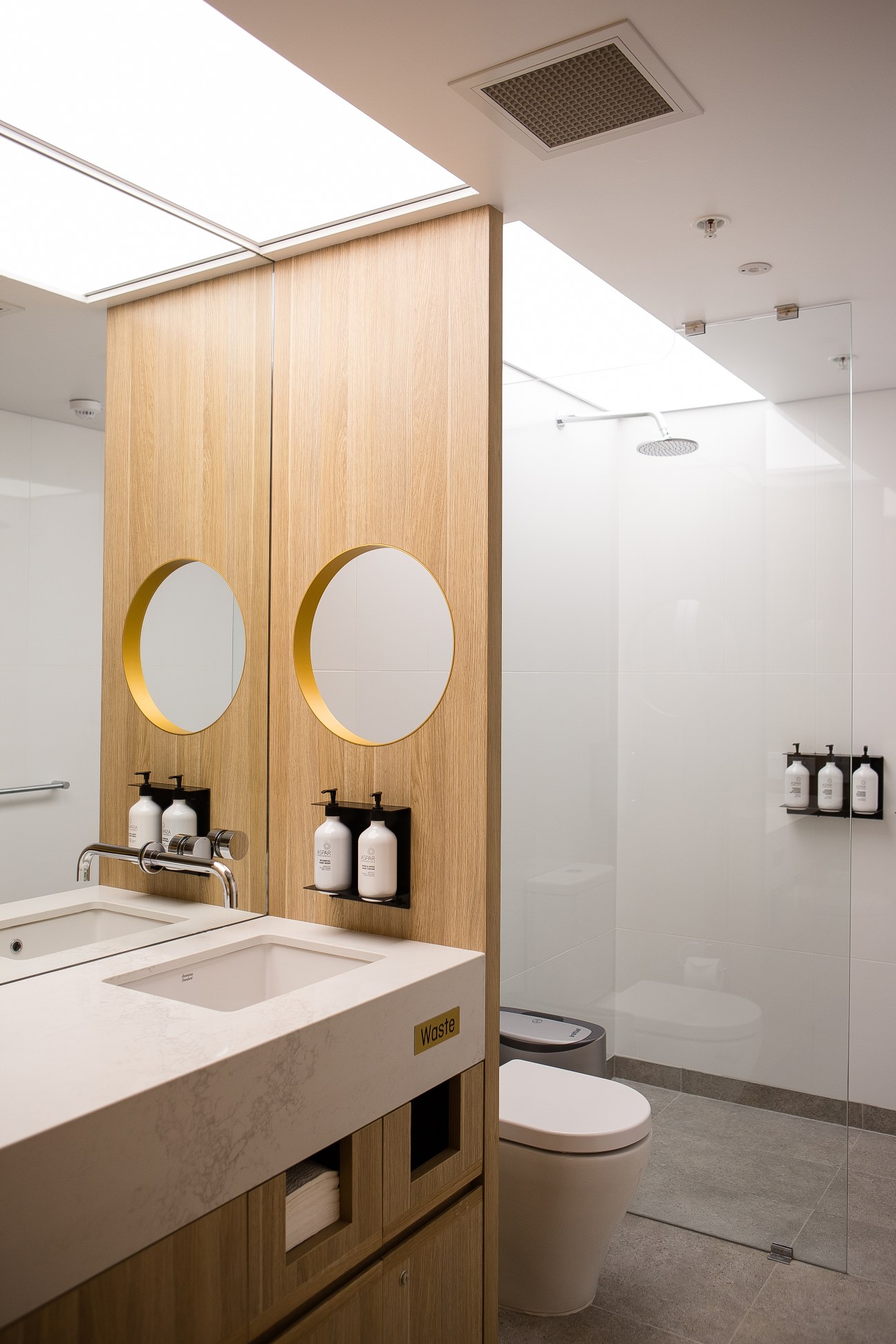 Modern bathroom with dual circular mirrors in wooden panels, marble sink, glass shower enclosure, and skylights above.