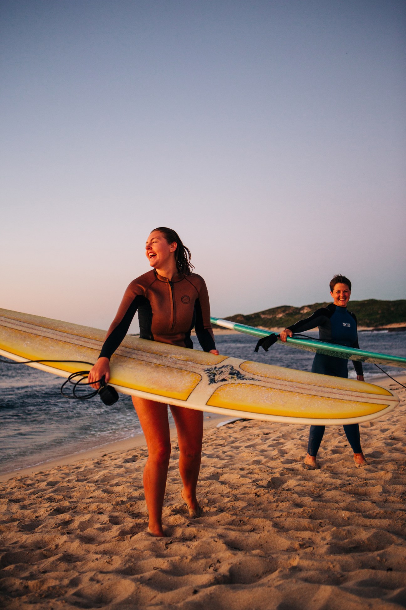 Two surfers in wetsuits carrying surfboards on beach at sunset