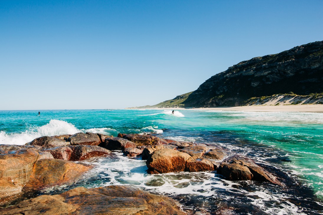 Coastal beach scene with turquoise waters, rocky foreground, sandy shore and hills in background