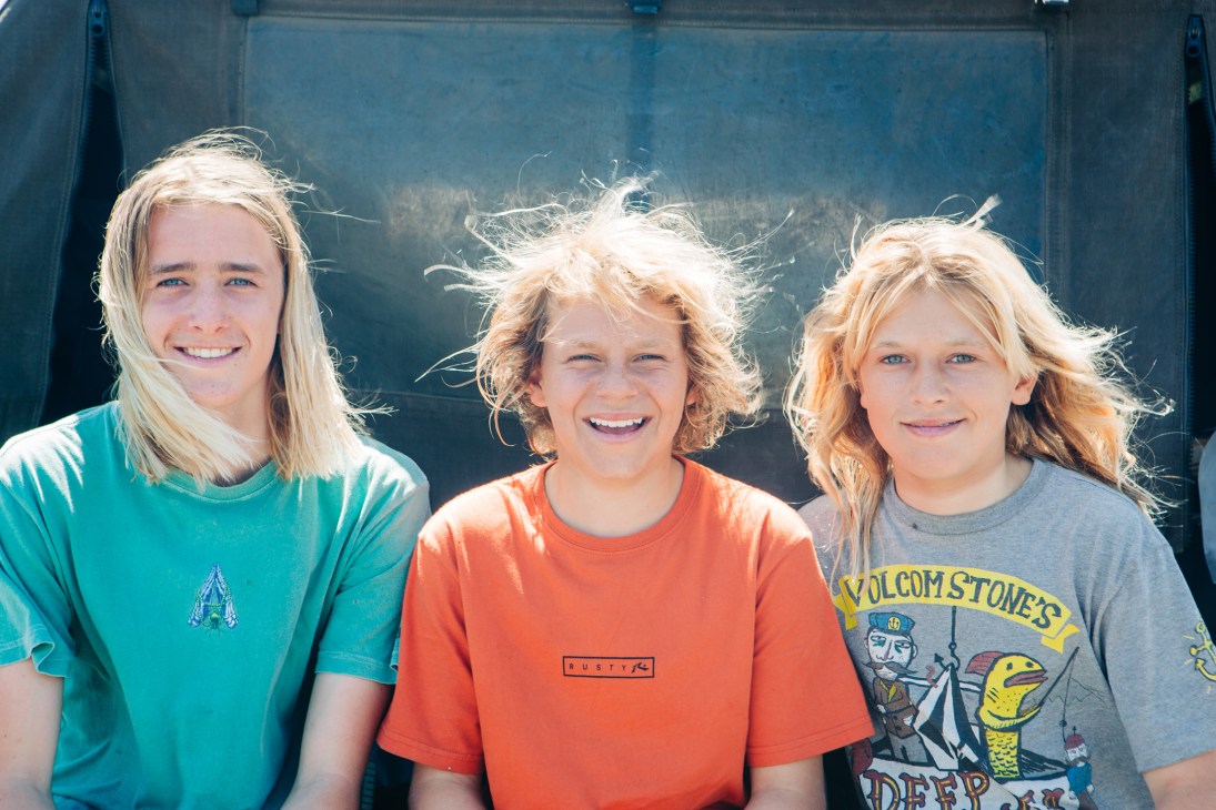 Three young blonde children standing together outdoors, wearing colorful t-shirts and smiling