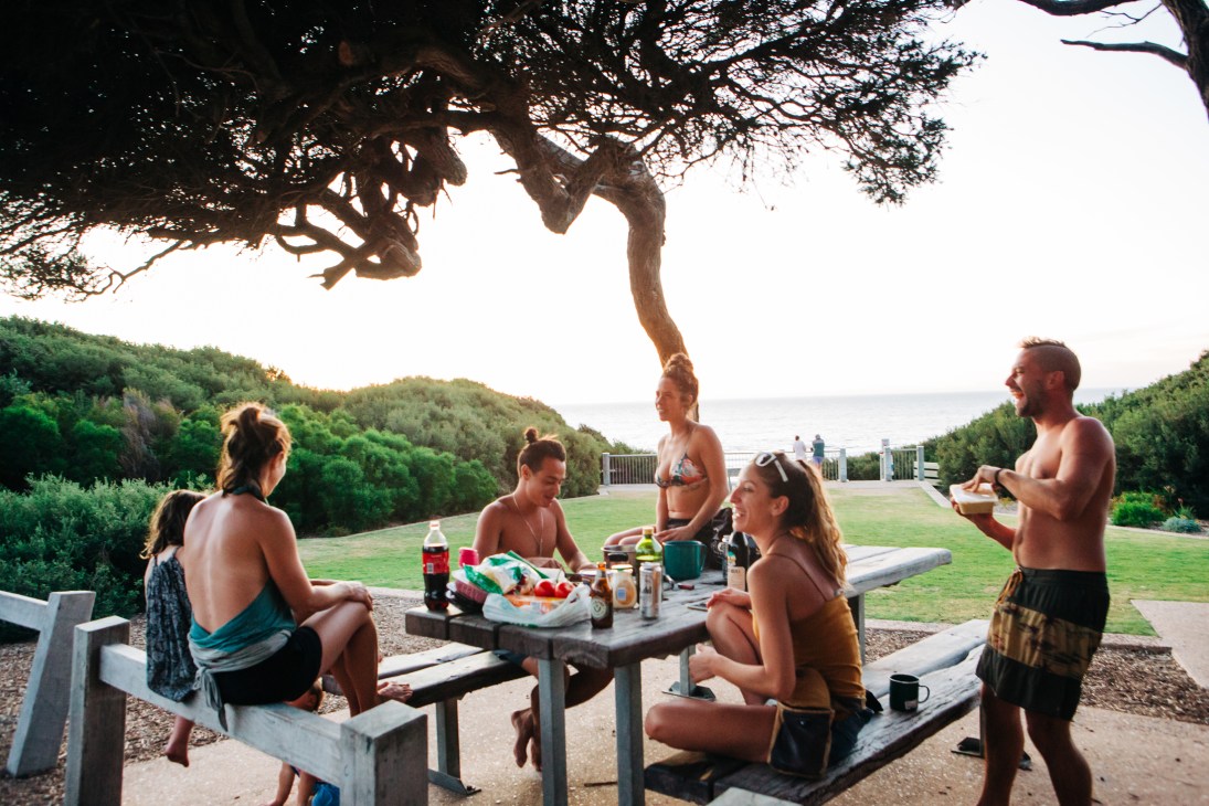 Group of travelers sitting around picnic table under tree at coastal camping area