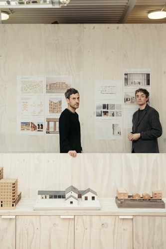 Two men standing behind a white table with architectural models and drawings on wall behind them