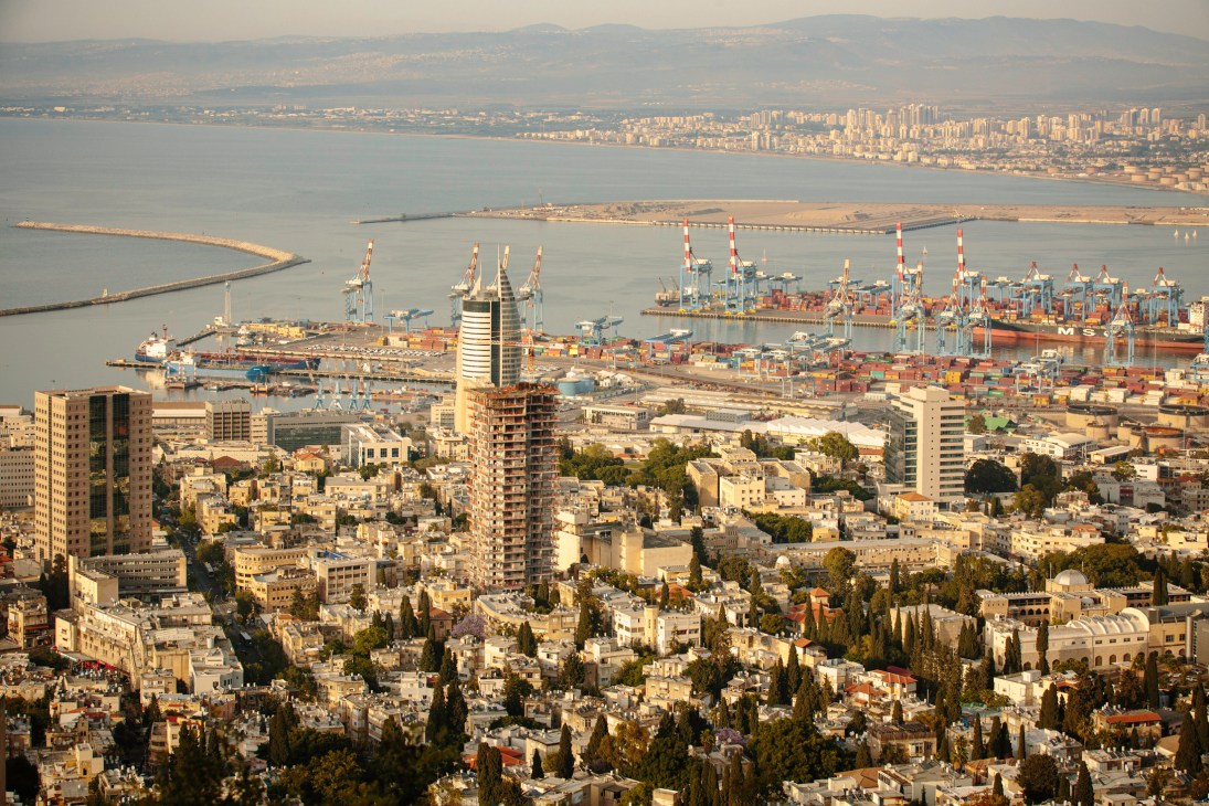 Aerial view of Haifa's port with cargo ships, cranes, and the city skyline beyond the Mediterranean Sea.