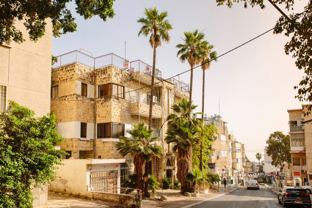 Palm-tree-lined street in Hadar HaCarmel showing Bauhaus villas with stone facades and rounded balconies.