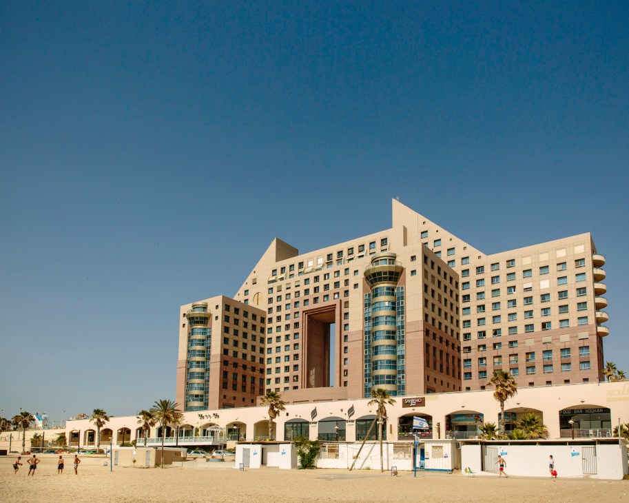 Postmodern beachfront building with cream and terracotta tones overlooking sandy beach with people and palm trees.