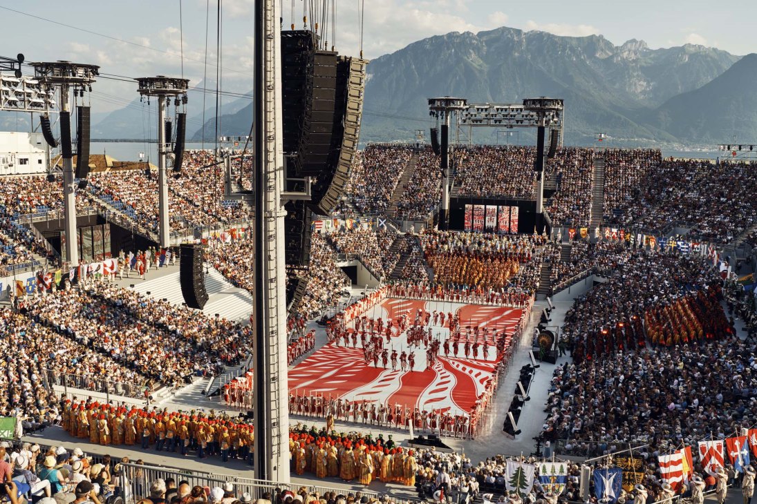 Aerial view of a temporary arena packed with thousands of spectators surrounding a central performance stage with mountains in the background.
