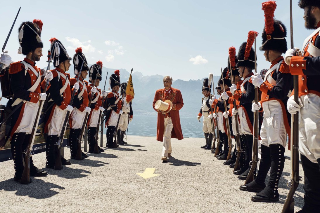 François Margot, Abbé Président, stands centered among costumed Swiss Guards holding rifles by Lake Geneva.