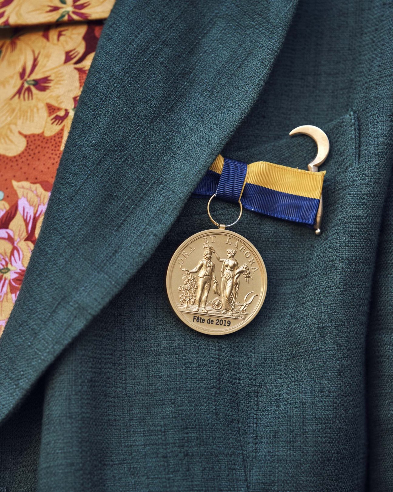 Gold medal with blue and gold ribbon on teal fabric, engraved 'Fête de 2019'