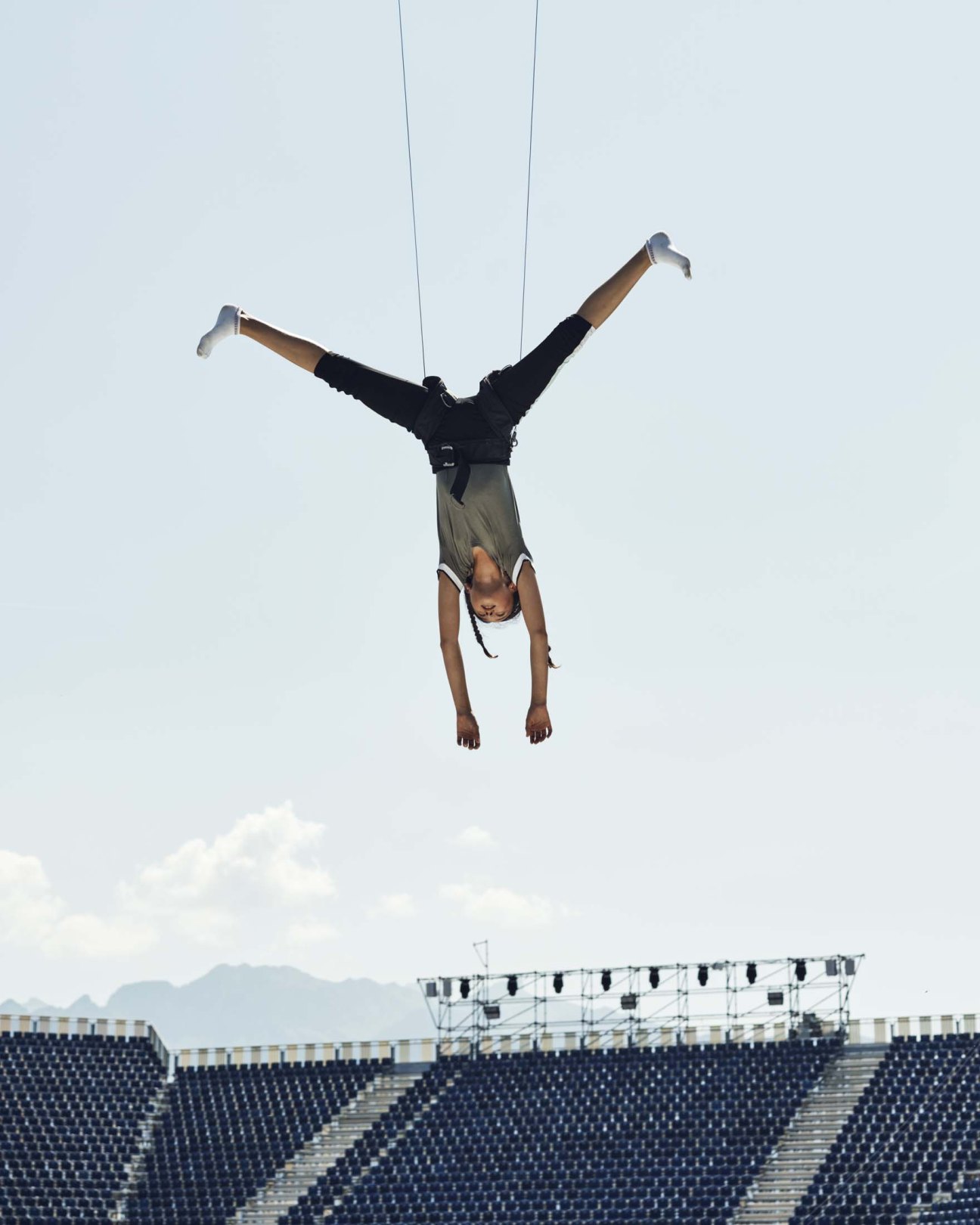Young woman performing aerial acrobatics suspended by ropes against sky with arena seating below