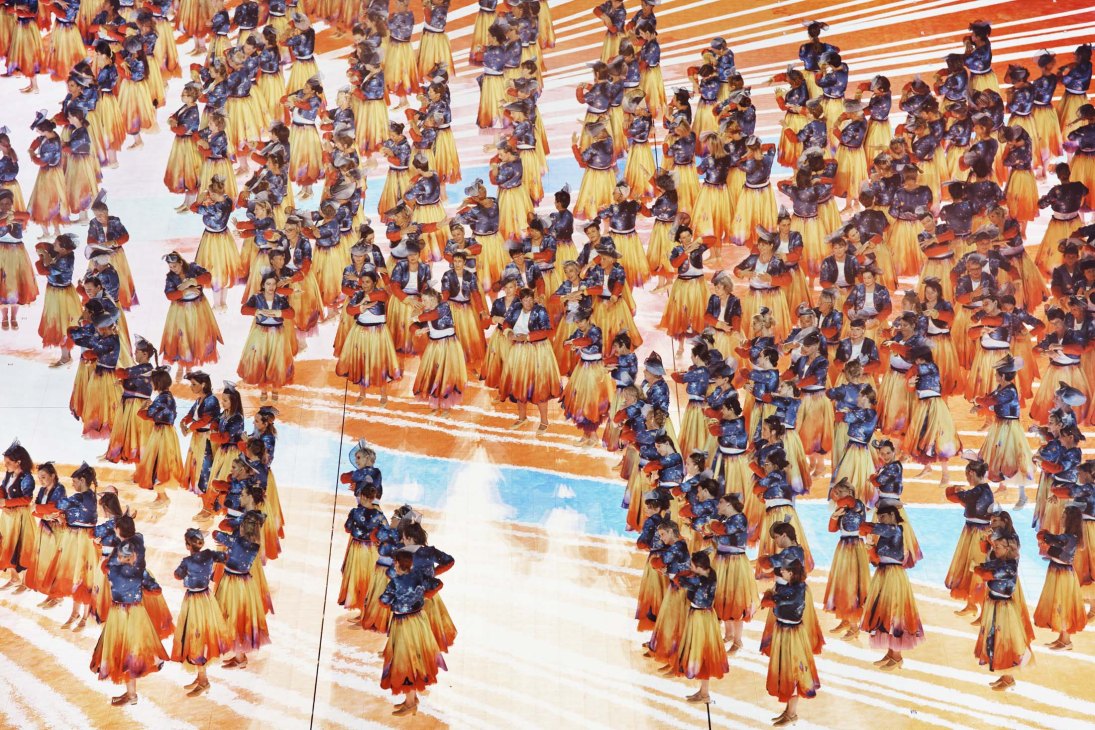 Aerial view of dozens of performers in golden-yellow and orange gradient skirts with blue tops on a striped stage floor.