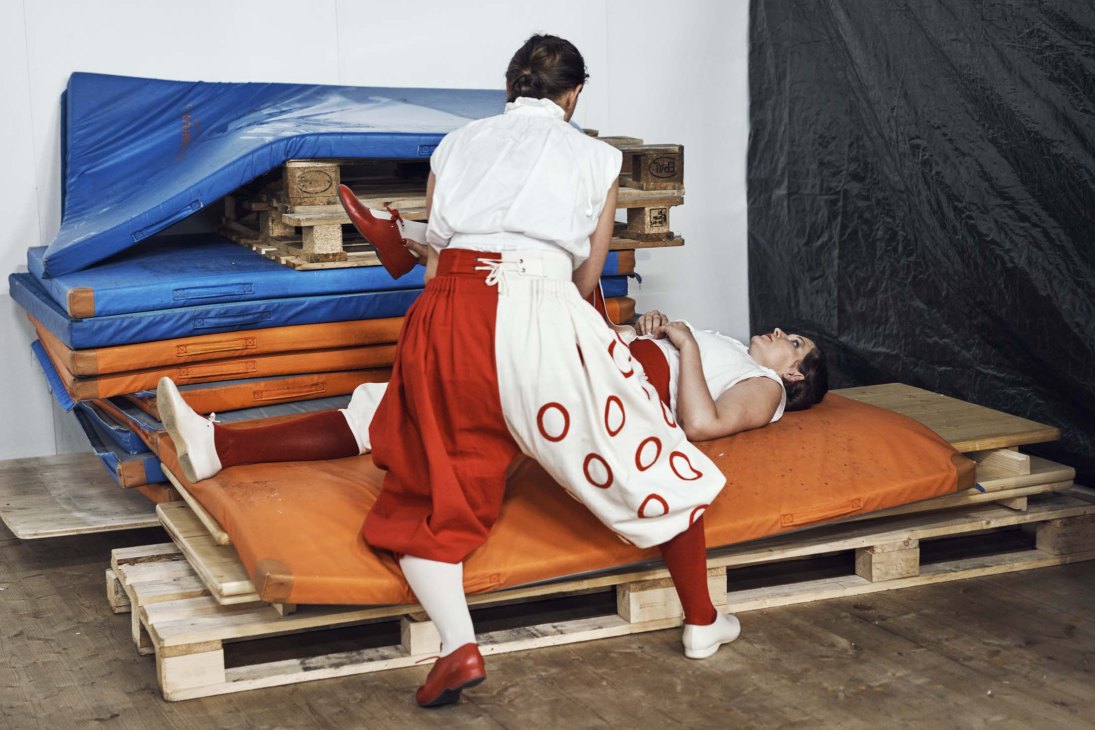 Performers in period breeches stretching on stacked colorful mattresses during festival rehearsal.