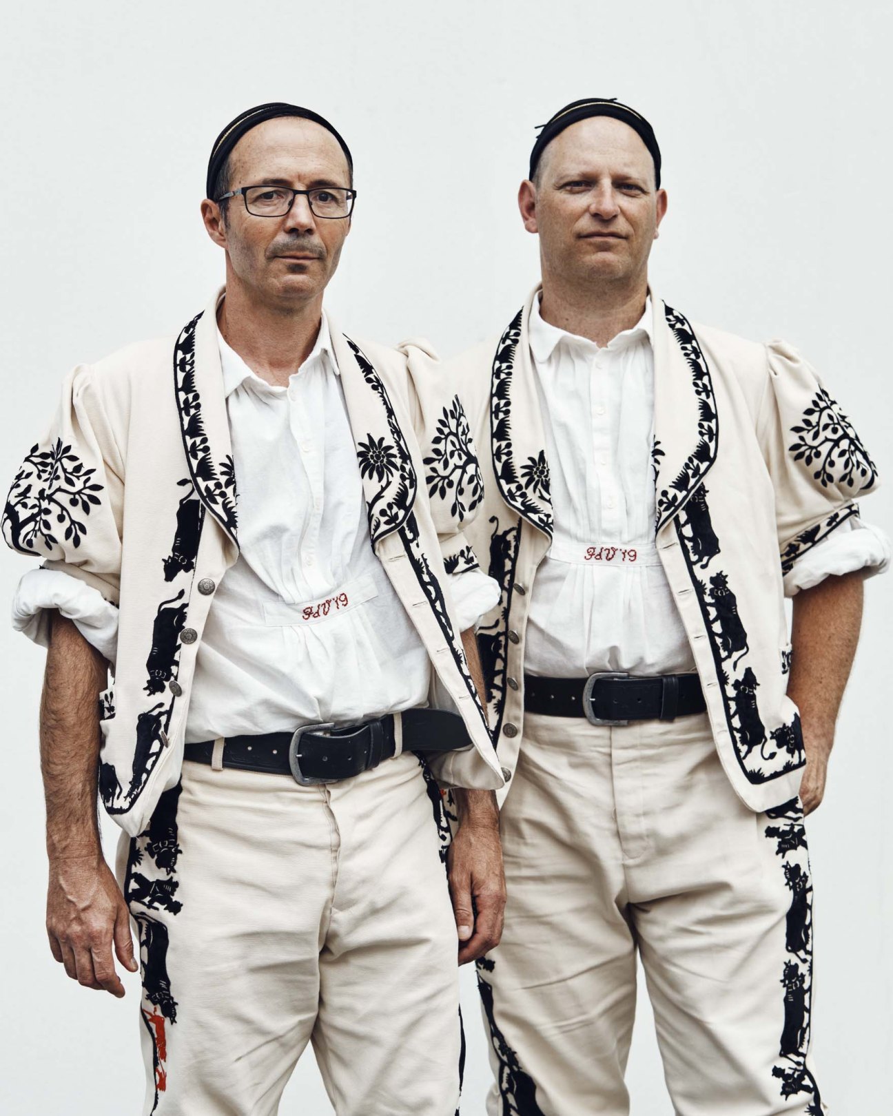 Two men in cream Alpine herdsman costumes with black and white embroidered vests and belts.