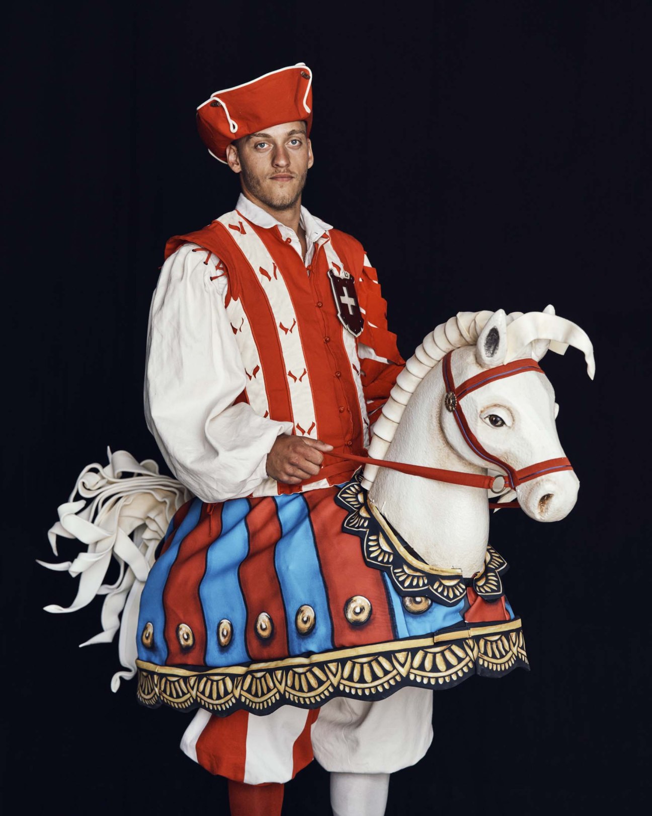 Man in red and white medieval knight costume holding horse reins beside white horse in ornate ceremonial tack.