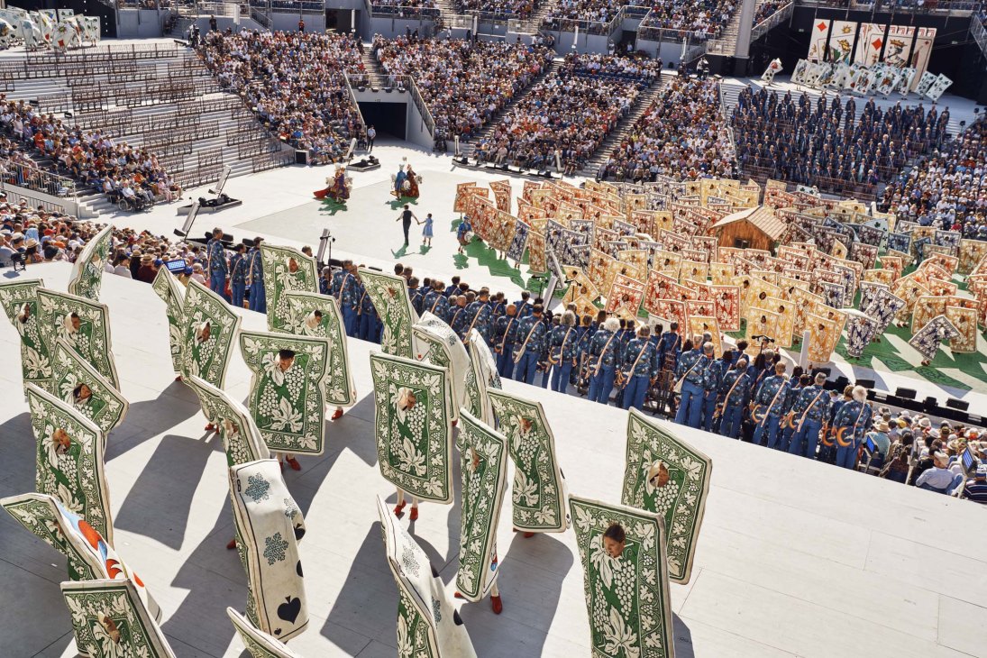 Aerial view of a packed arena with thousands of spectators watching a theatrical performance with costumed performers on stage.