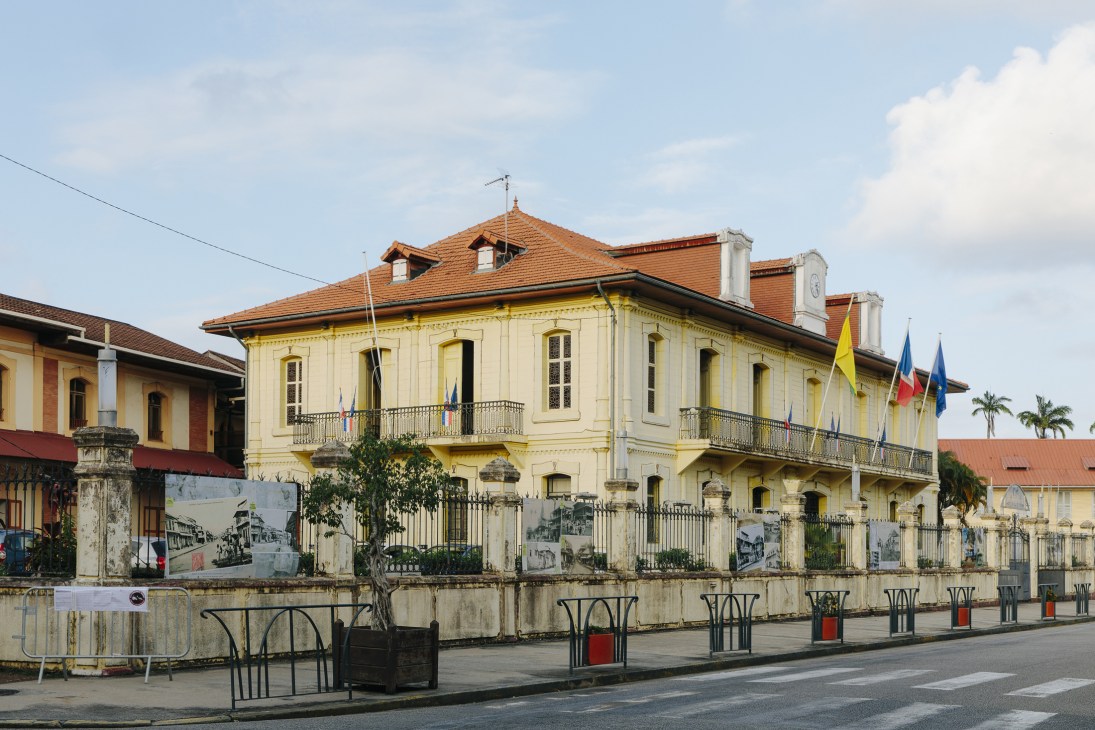Pale yellow colonial-style town hall building with terracotta roof in Cayenne, French Guiana, featuring ornate balconies and flags.