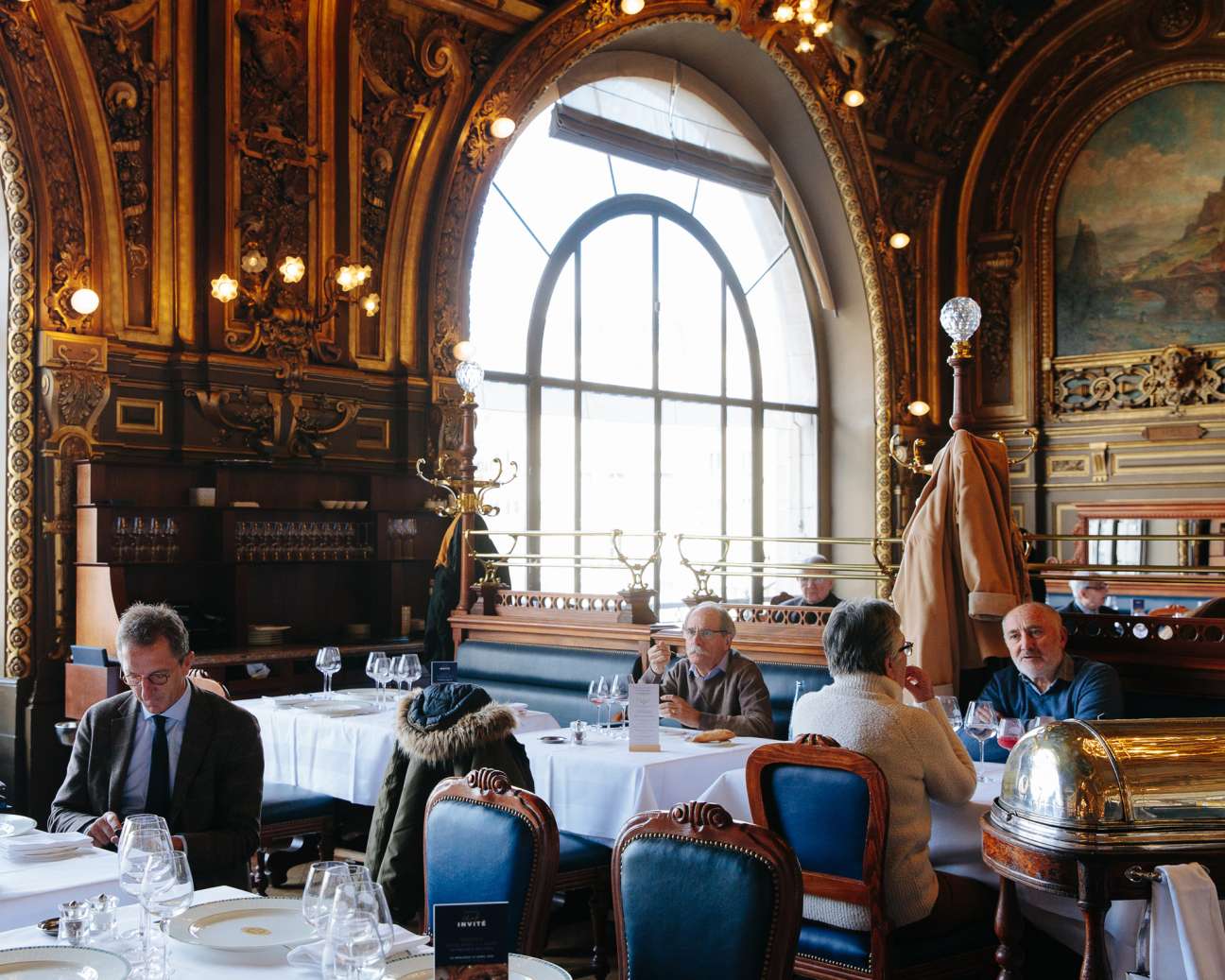 Diners seated at white-clothed tables in Le Train Bleu's ornate gilded dining room with arched windows.
