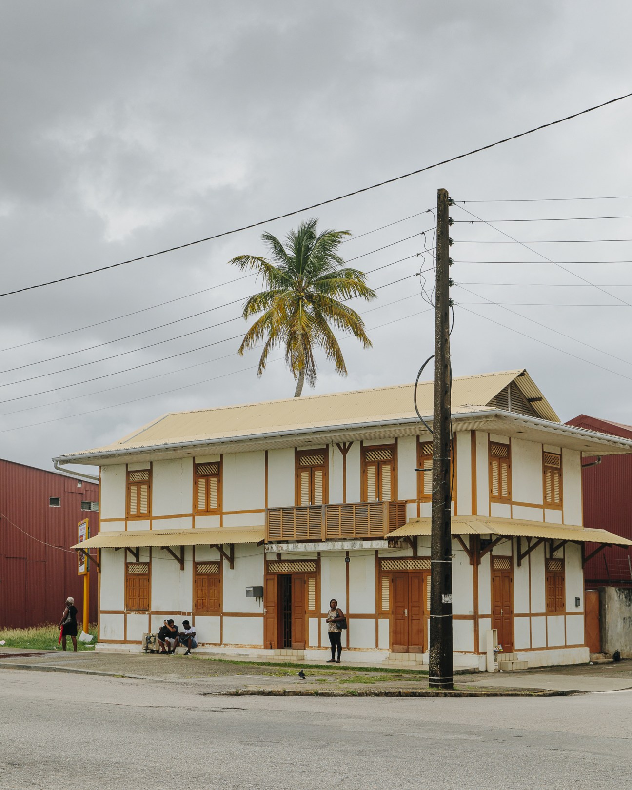 White Creole-style two-story building with wooden shutters and a palm tree in front under cloudy sky in Cayenne, French Guiana.
