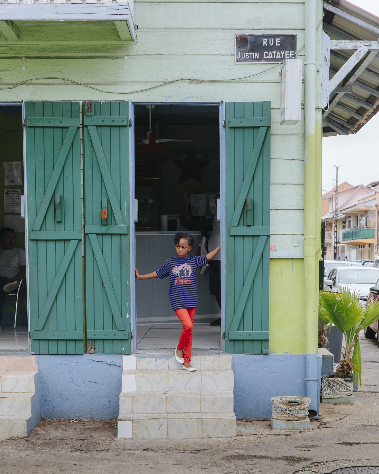 Child in striped shirt and red leggings standing in doorway of Creole-style building with green shutters in Cayenne.