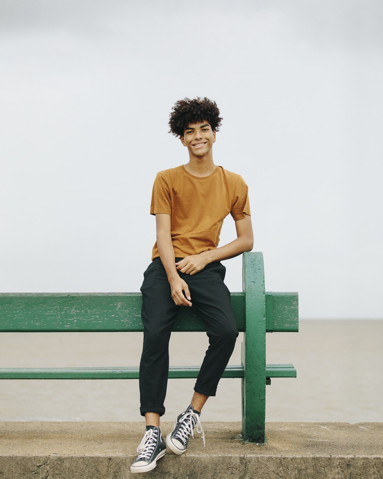 Young man in mustard t-shirt and black pants sitting on green bench.