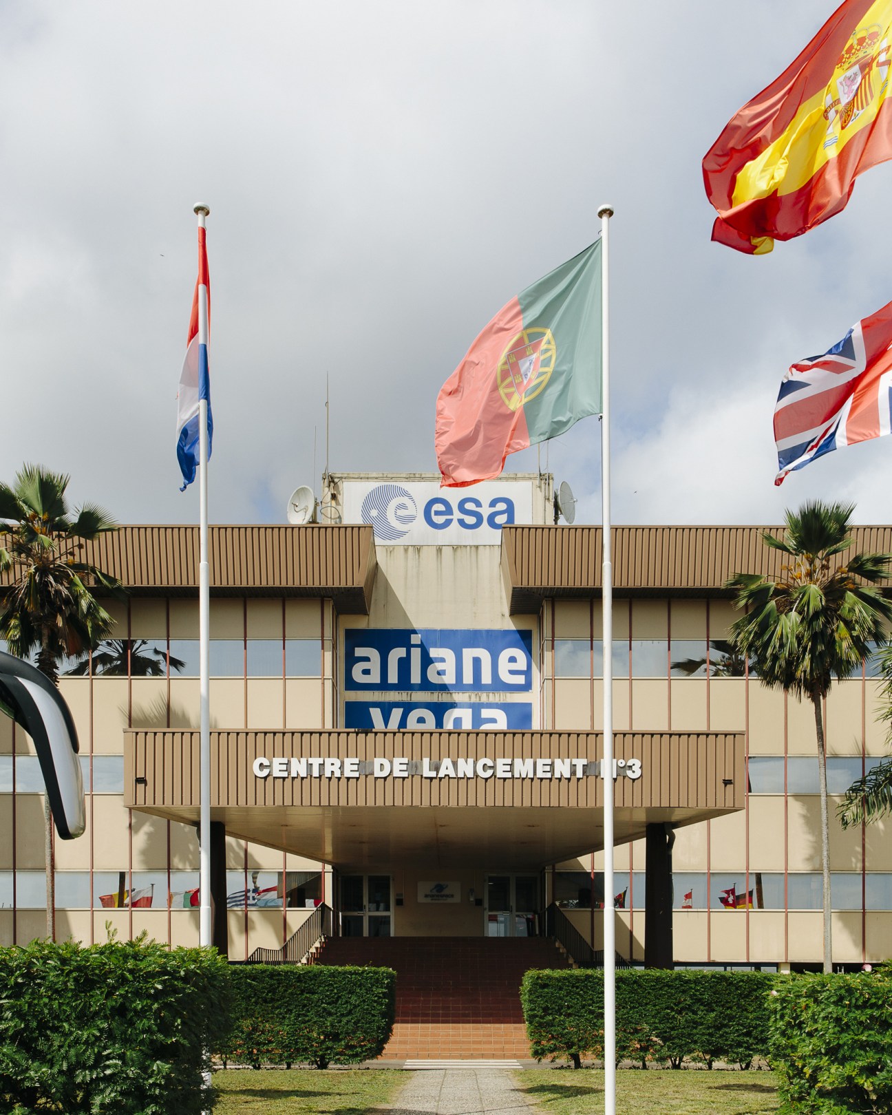 Kourou Space Centre building with ESA and Ariane signage, palm trees, and international flags flying outside.