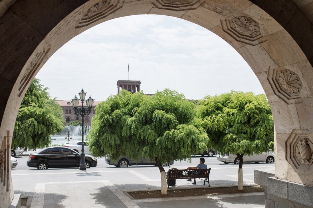 A man sits on a bench under a stone archway with ornate carvings, overlooking Republic Square in Yerevan with green trees and government buildings.