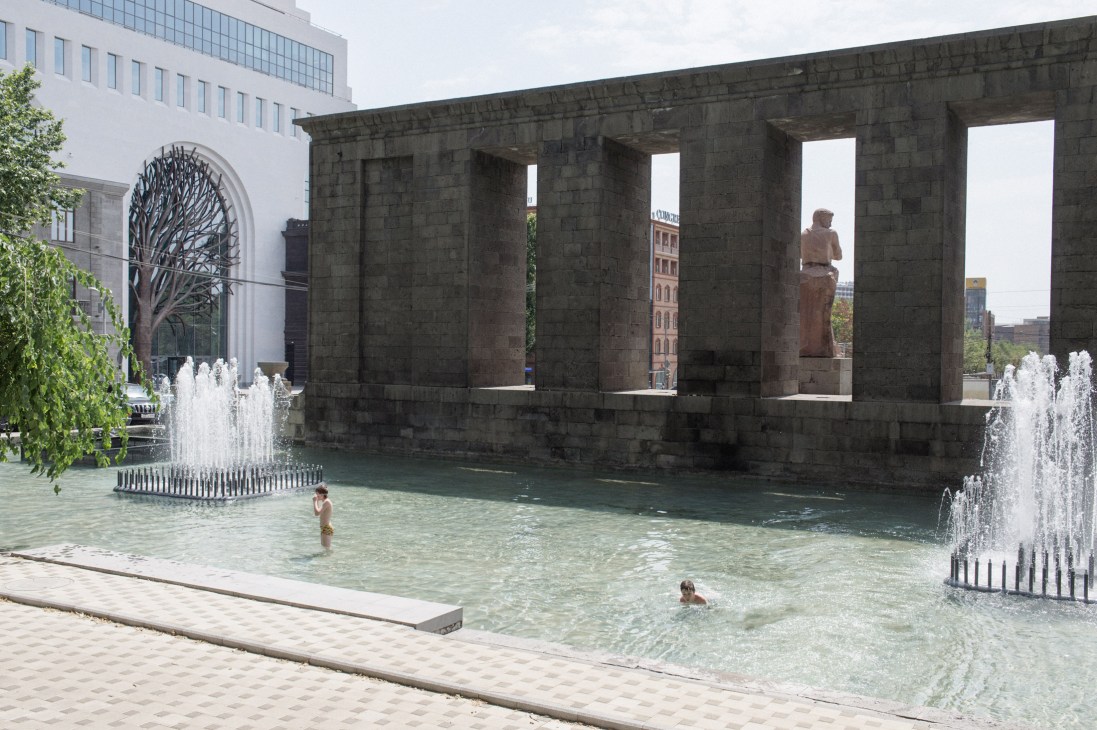 People swimming in a turquoise pool beside a massive stone monument structure with fountains.