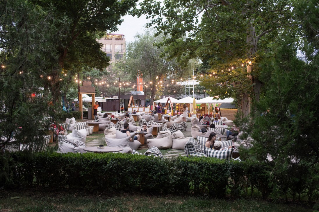 People relaxing on striped cushions and bean bags at an outdoor café surrounded by trees with string lights and vendor stalls in the background.
