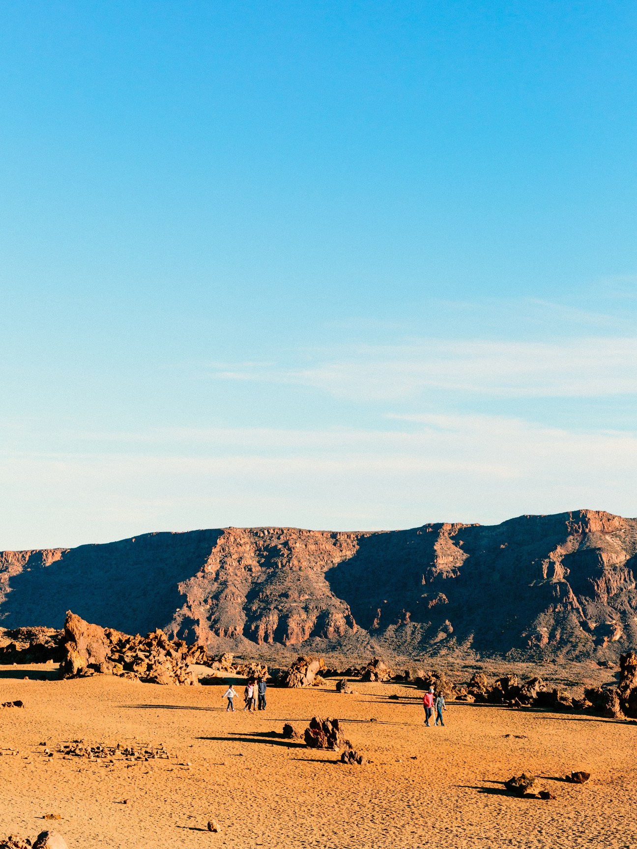 Visitors walking across golden volcanic sand with dramatic rocky cliffs rising against blue sky in Tenerife.