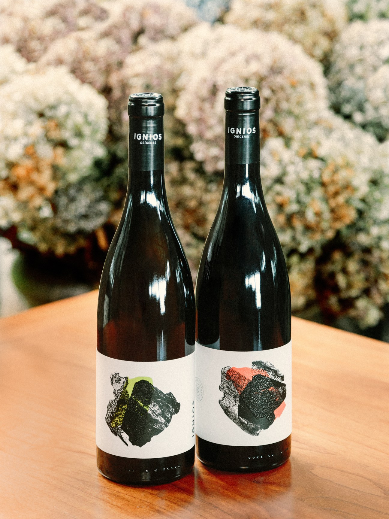 Two Ignios Orígenes wine bottles with artistic label designs displayed on a wooden surface against flowering foliage.