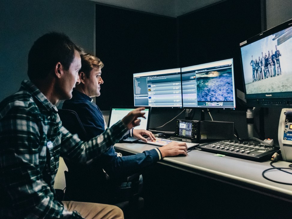 Two men work at a control room desk with multiple monitors displaying broadcast content and data.