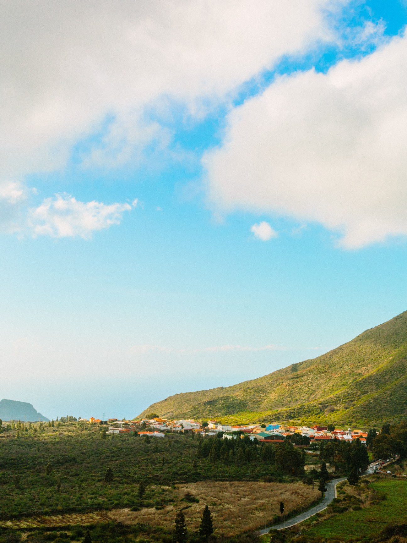 A hillside village with colorful houses nestled against a green mountainside under blue sky with clouds.