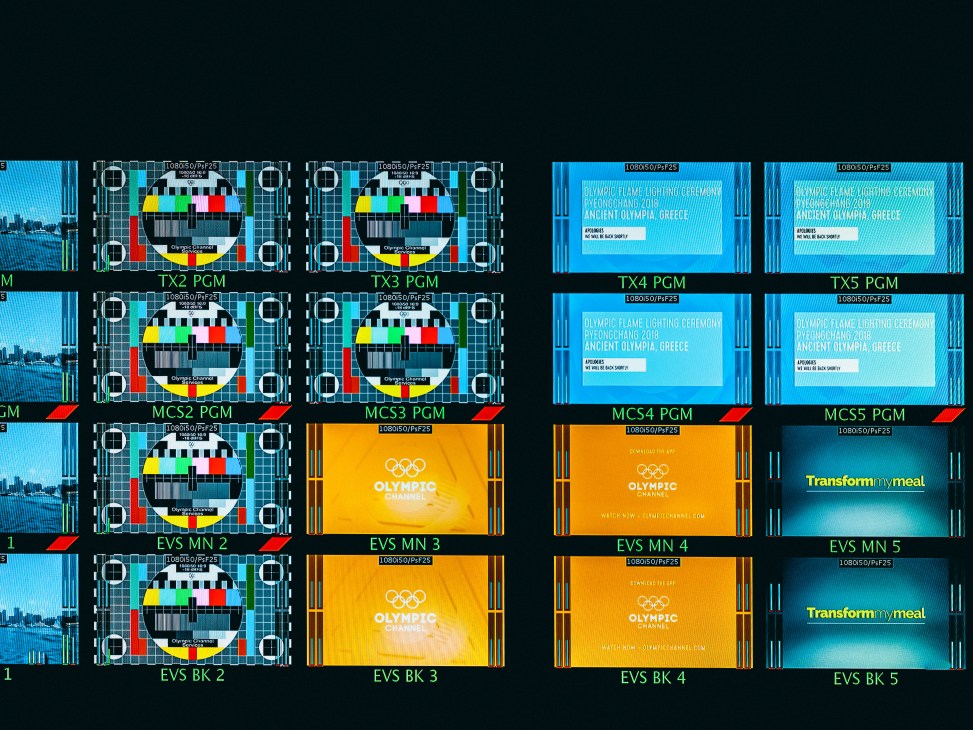 Multiple broadcast monitor screens displaying test patterns, Olympic Channel branding, and program content in a control room.