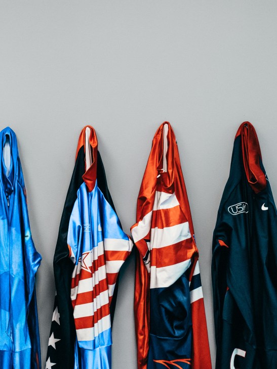 Four folded Olympic team jackets in blue, red, white and navy hanging against a grey wall.
