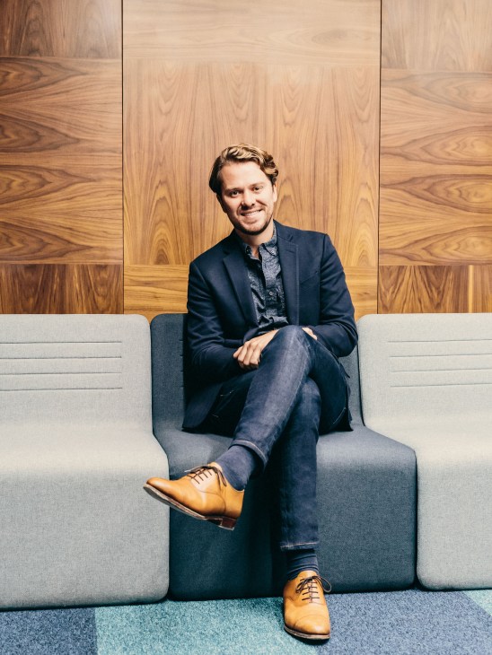 Greg Groggel, director of original programming, sitting on a gray sofa against a wooden panel backdrop.