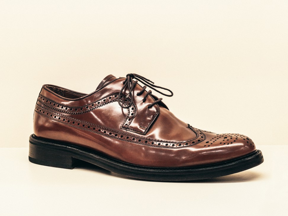 Brown leather wingtip brogue with black laces and perforated detailing on a cream background.