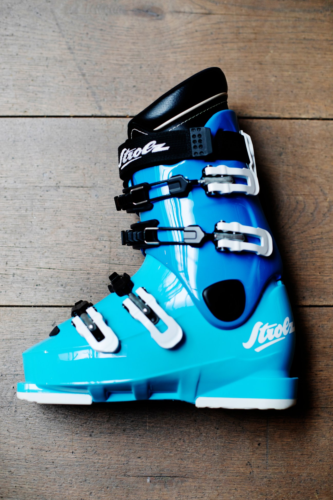 A bright blue Strolz ski boot with black and white buckles on a wooden floor.
