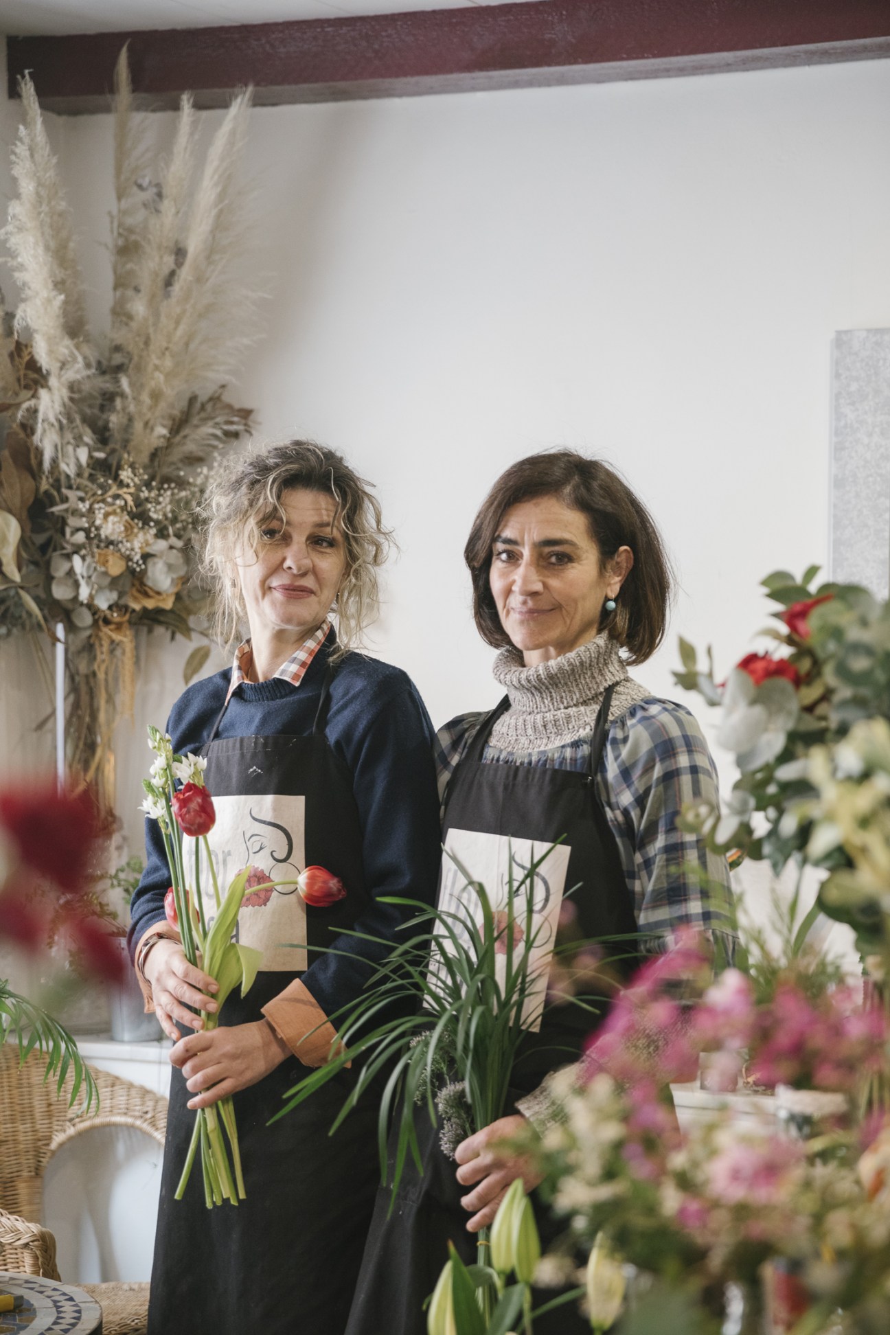 Two women at Flor de Lola flower shop holding bouquets surrounded by fresh flowers and plants.