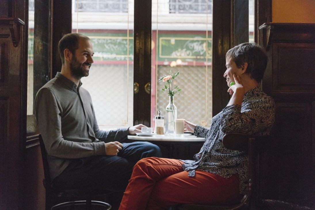 Miguel Oyarzun and Isla Aguilar sit at a café table by a window with decorative glass panels, smiling while in conversation.
