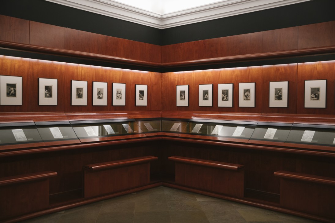 Wood-paneled gallery room displaying framed Goya engravings on burgundy walls with display cases below.