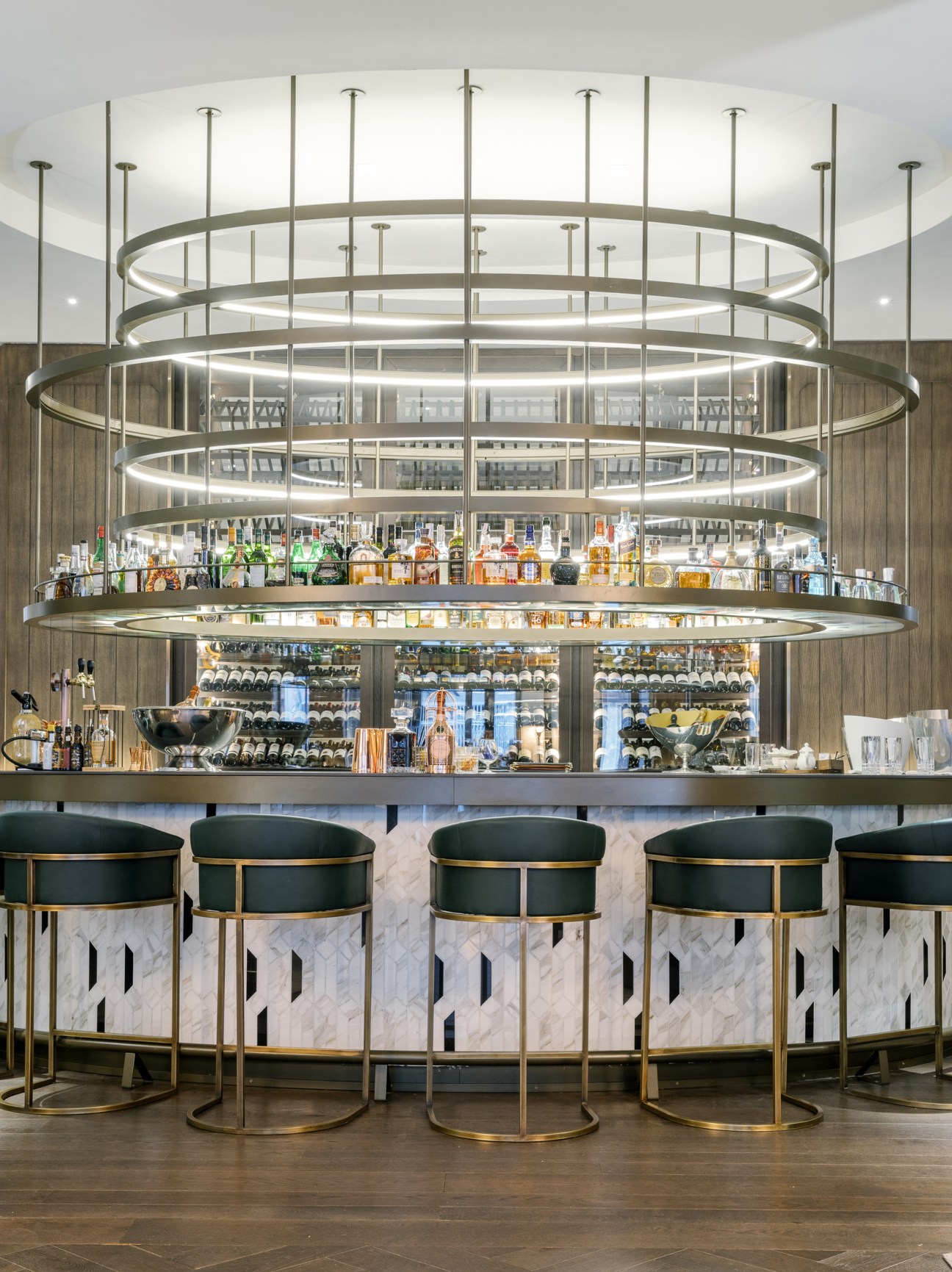 Modern circular bar with multiple tiers of concentric rings lit with LED strips, stocked with spirits and bottles, featuring black leather and brass bar stools.