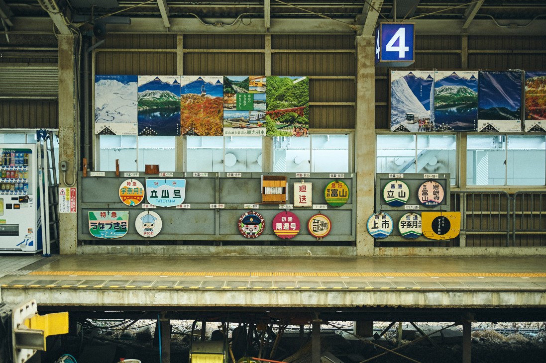 Japanese railway station platform with mountain landscape posters and colorful destination signs displayed on concrete wall.