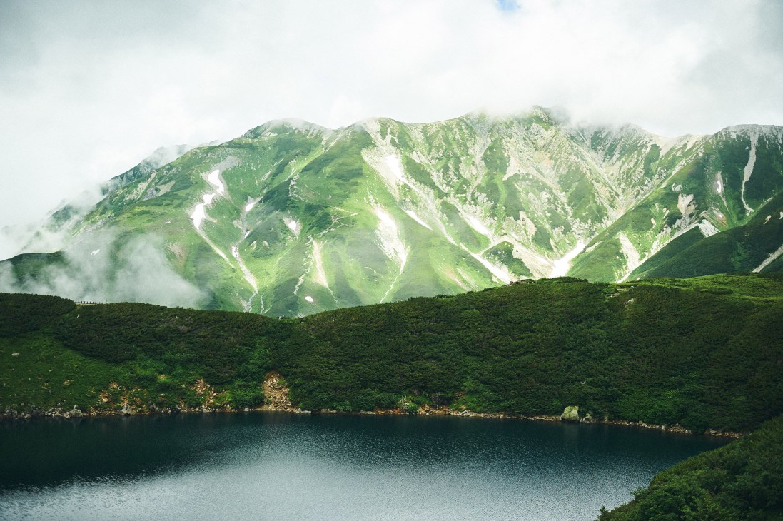 Tateyama mountain range with verdant valleys, white rocky slopes, and mist-covered peaks rising above a dark forested shoreline and alpine lake.