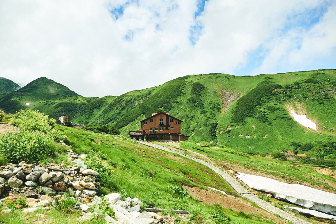 Mountain lodge nestled in lush green valley surrounded by verdant peaks in Japan's Tateyama range.